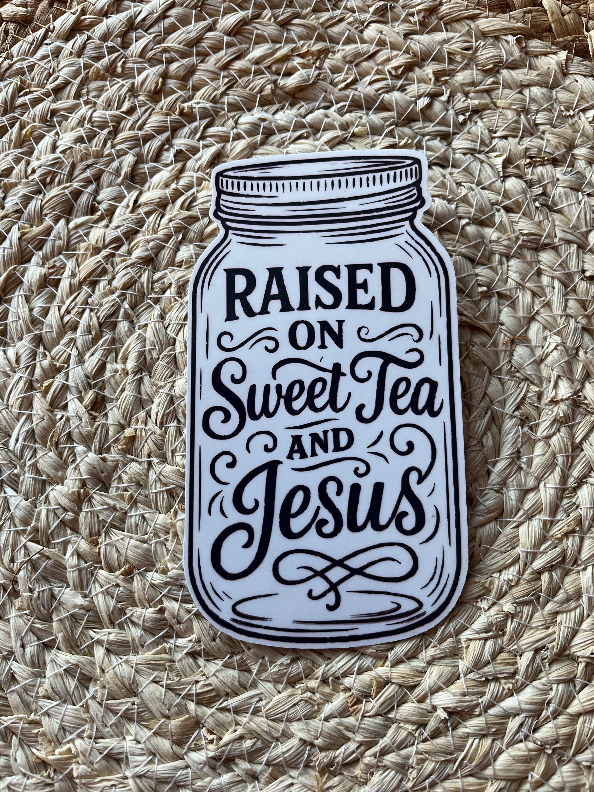 Sticker of a mason jar with text 'Raised on Sweet Tea and Jesus' on a woven mat.