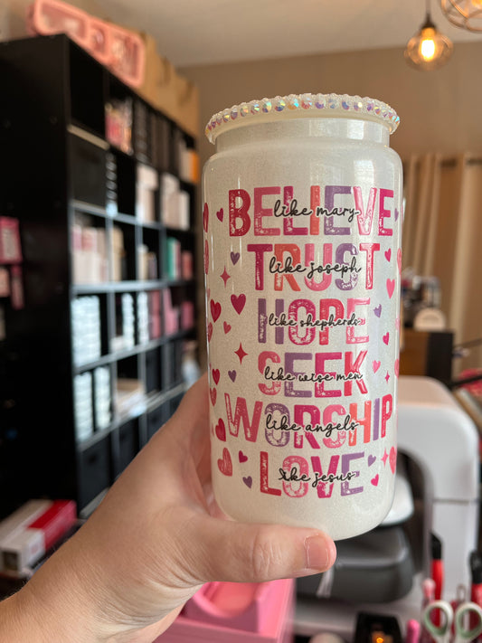 Hand holding a white tumbler with motivational phrases and rhinestones, against a blurred background of makeup products.