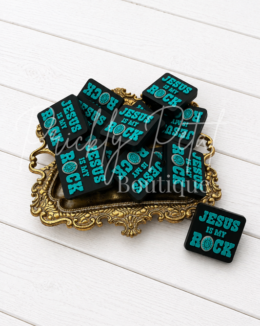 silicone focal bead with turquoise text reading 'JESUS IS MY ROCK' arranged on an ornate gold tray, with one pin placed beside it on a white wood plank background. 
