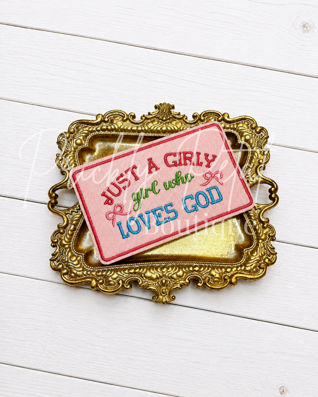 A rectangular embroidered patch with the phrase 'JUST A GIRLY girl who LOVES GOD' in red, green, and blue lettering on a pink background, displayed on an ornate gold picture frame over a white wood plank surface.