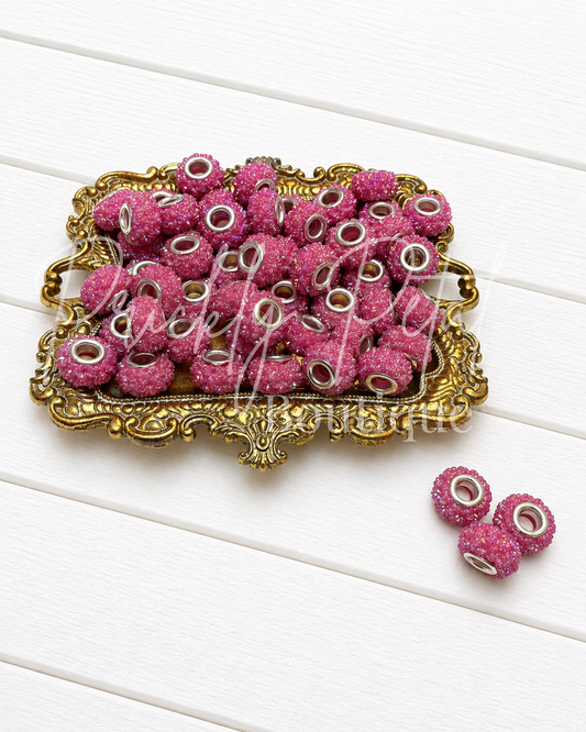 Pink glittery round beads with white and silver center details, displayed in an ornate gold tray on a white wood background.