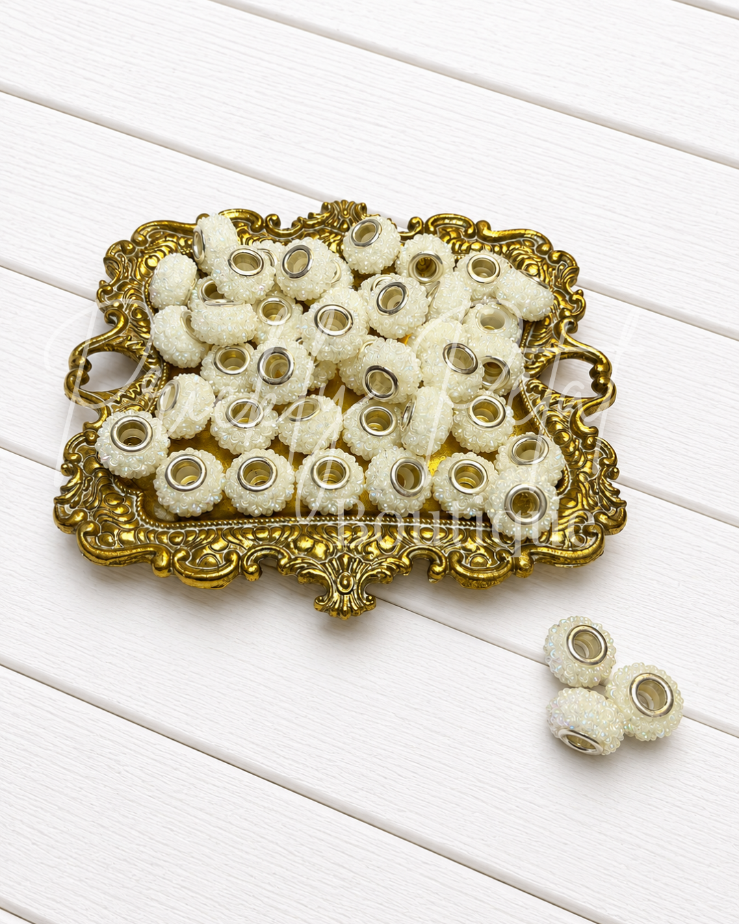 A collection of small white pom-pom beads with gold metal cores, displayed on an ornate gold-colored tray with three additional beads placed beside it on a white wooden background.