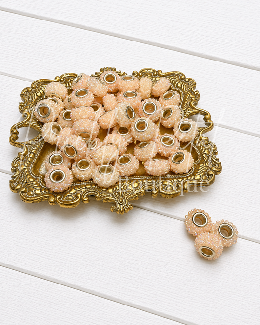 A pile of peach-colored textured beads with gold-toned metal centers, displayed on an ornate gold tray with three additional beads placed beside it on a white wooden surface.