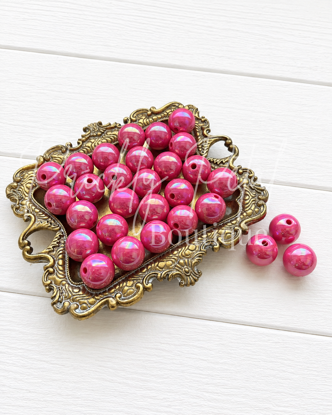 Bright pink iridescent round craft beads with holes, displayed in an ornate gold-colored metal dish on a white wood background.