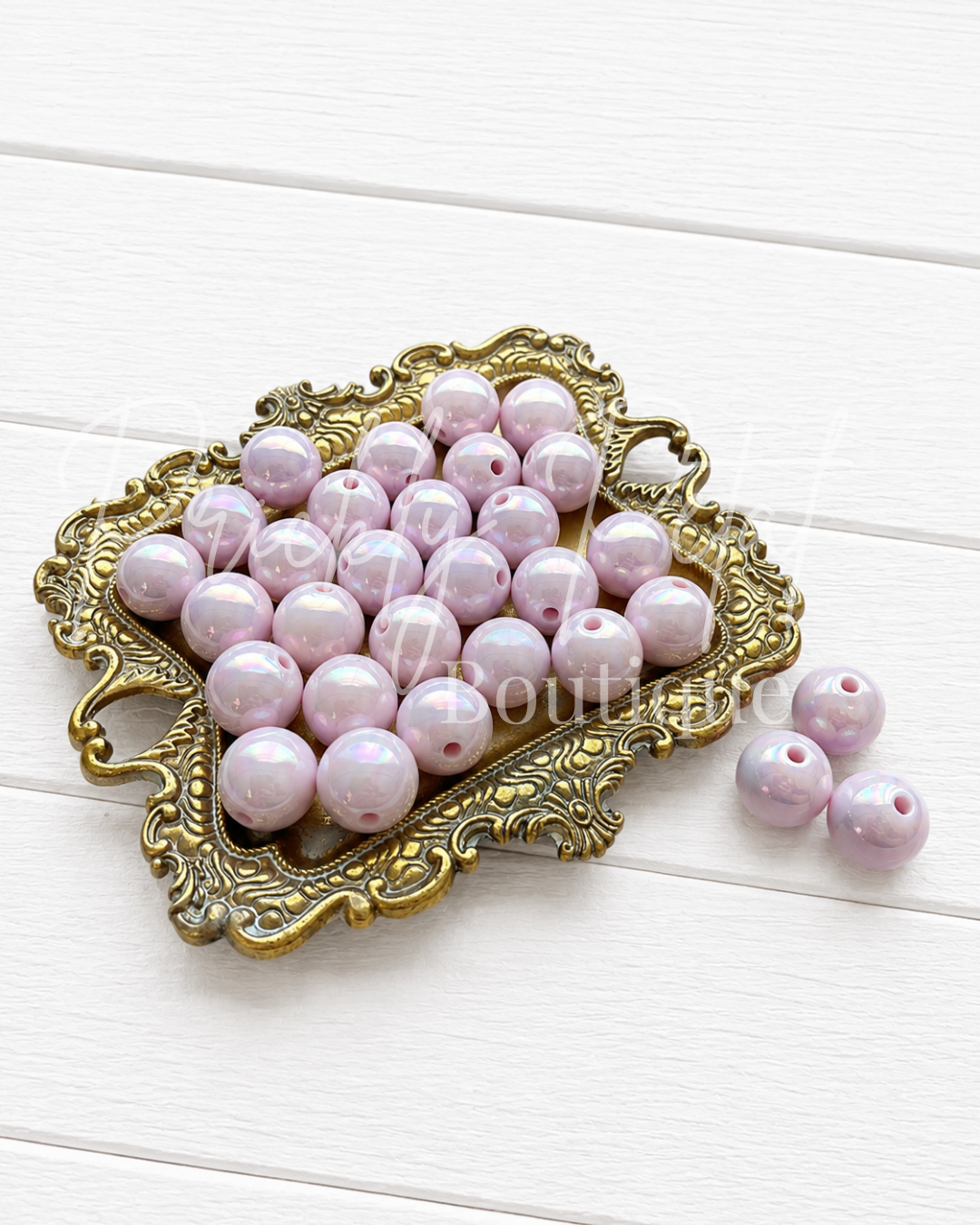 Light pink round craft beads with a glossy finish, displayed on an ornate gold-colored metal tray with three additional beads placed beside it on a white wooden background.
