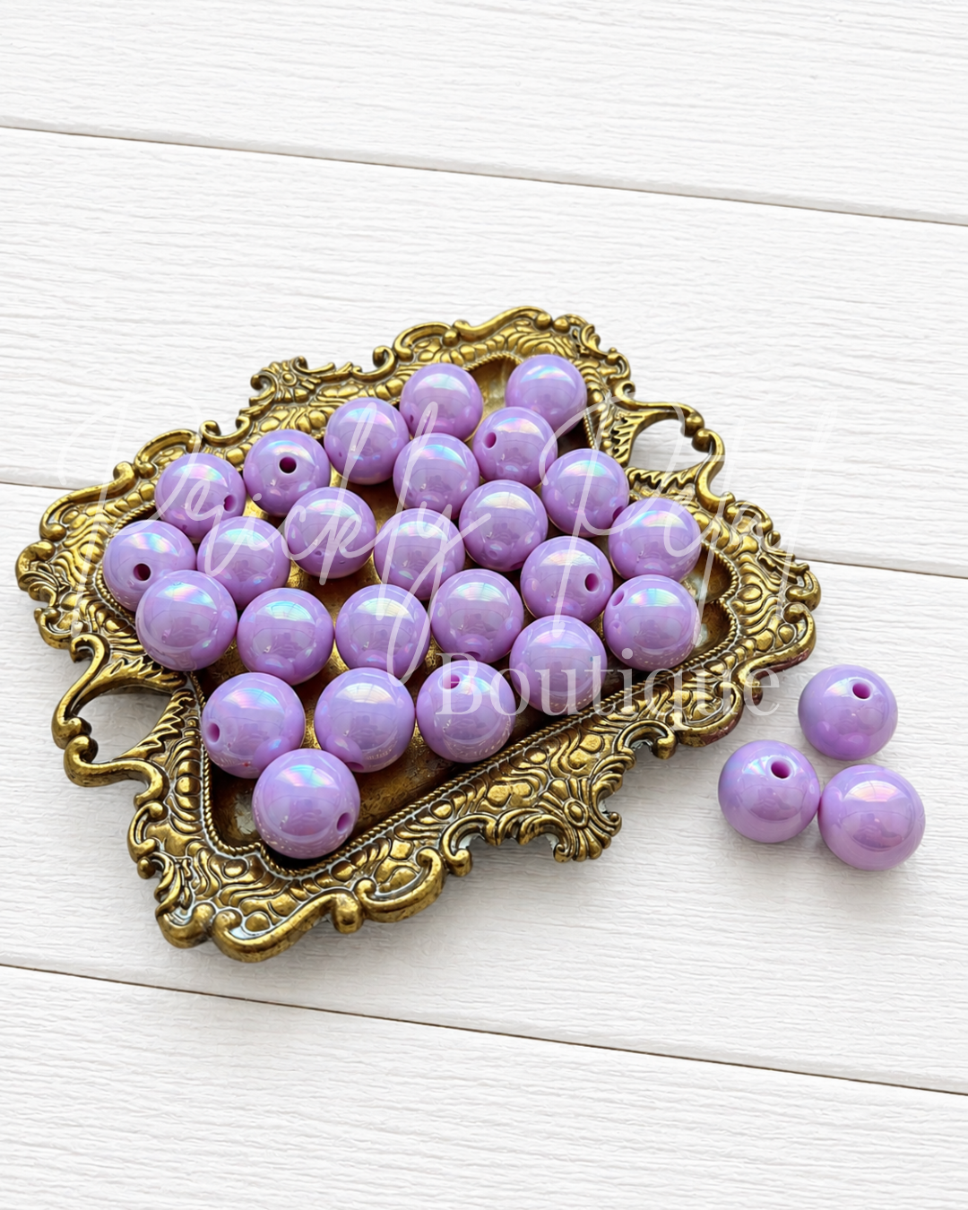 Purple round craft beads with a glossy finish, displayed on an ornate gold-colored metal tray with three additional beads placed beside it on a white wooden background.
