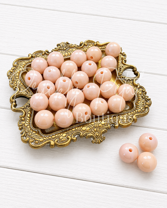 Light peach-pink round craft beads with center holes, displayed in an ornate gold tray with three additional beads on a white wood background.