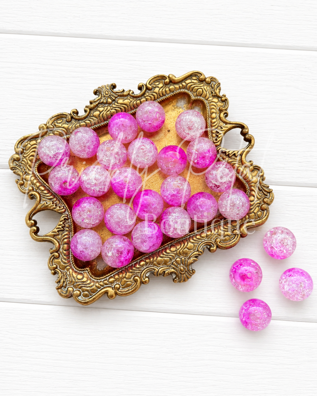 Pink and white glittery round beads displayed in an ornate gold tray on a white wooden background, with three loose beads to the right.