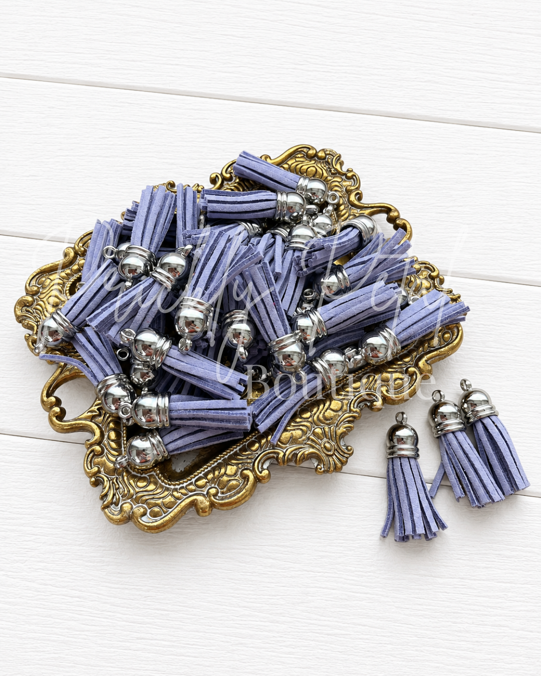 Lavender purple tassel charms with silver-toned metal caps, displayed on an ornate gold tray with two matching tassel earrings on a white wood background.