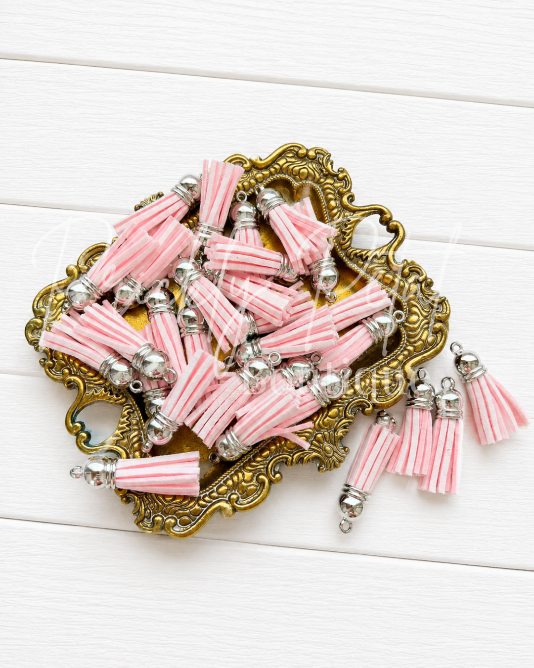 Pink and white striped tassel charms with silver metal caps, displayed on an ornate gold tray over a white wood background.