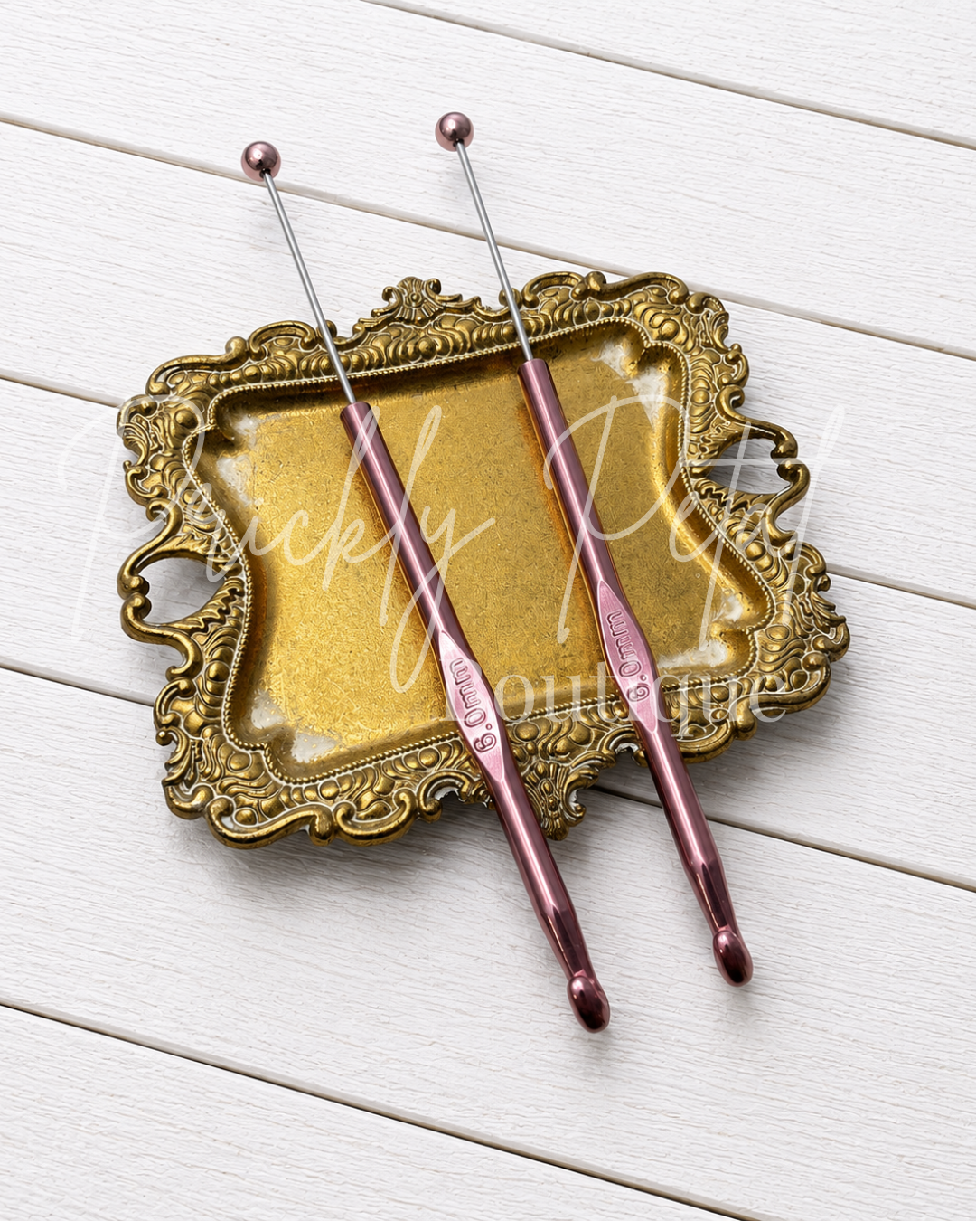 Two metallic crochet hooks with a rose gold finish resting on an ornate gold-colored decorative tray, set against a white wood plank background.