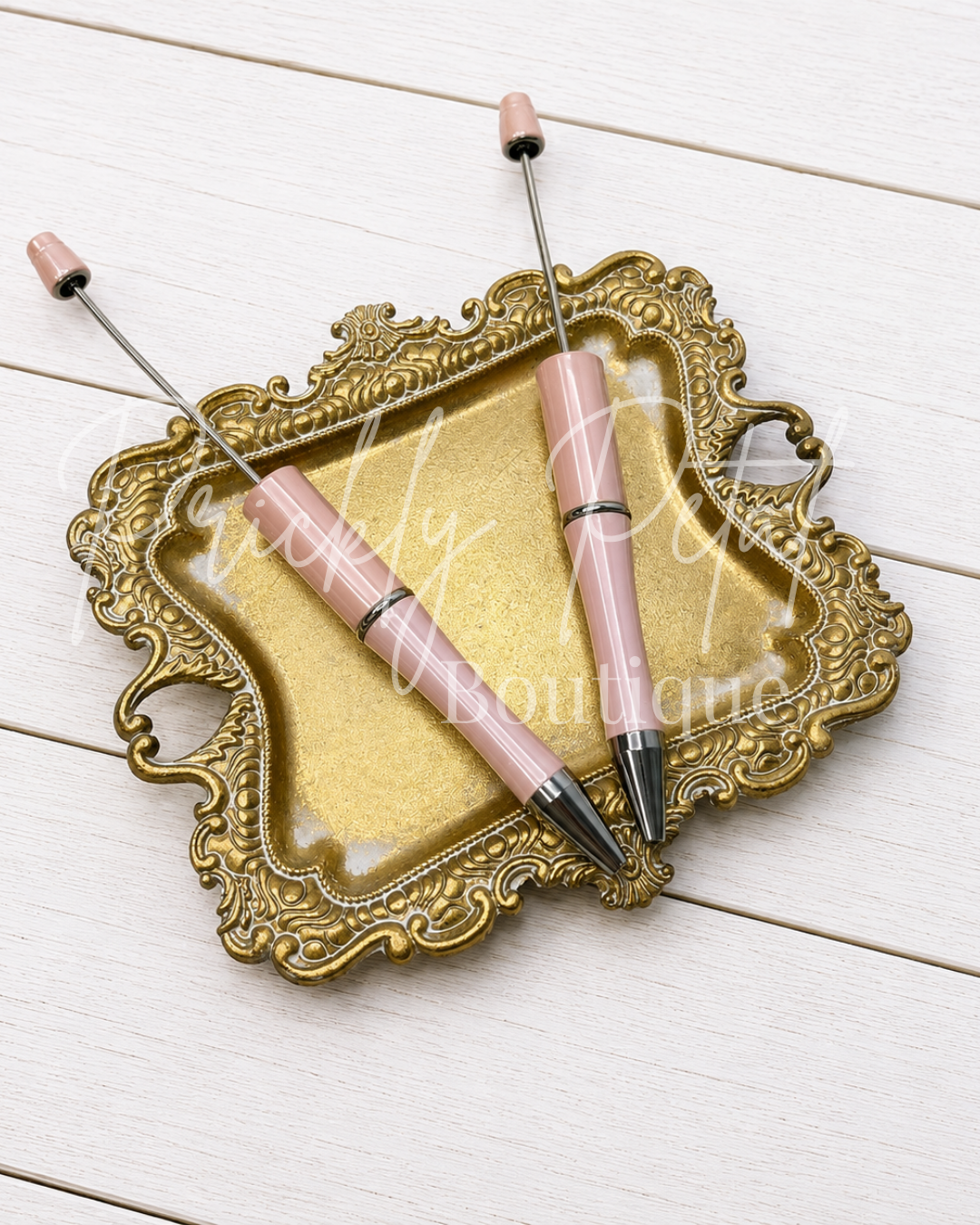 Two pink and silver ballpoint pens resting on an ornate gold-colored metal tray, placed on a white wooden background.