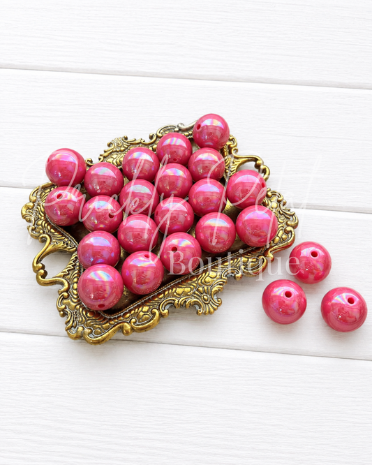 A pile of glossy pink round beads with a small hole, displayed on an ornate gold-colored metal tray with three beads placed beside it on a white wood background.
