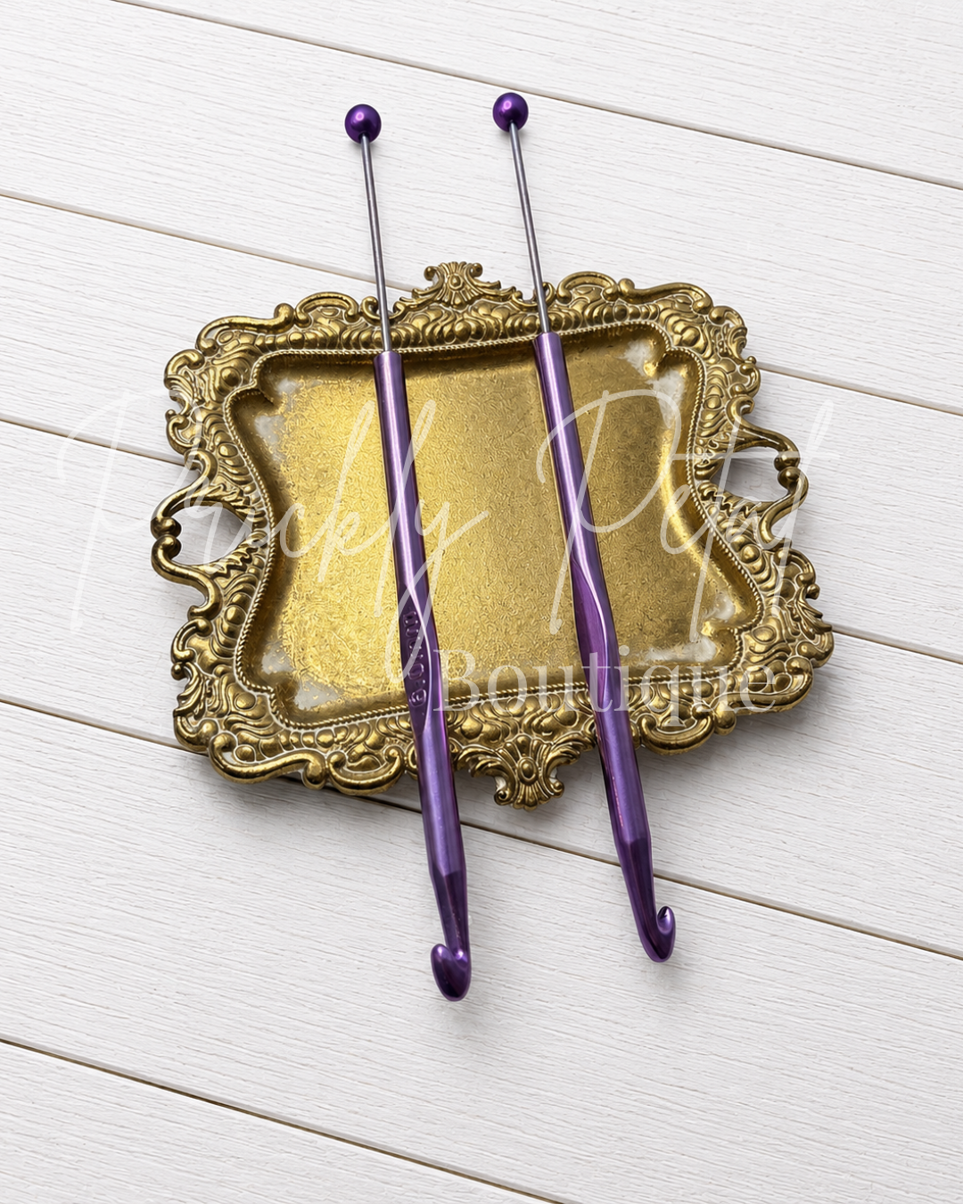 Two purple crochet hooks with silver tips and ball ends, displayed on an ornate gold tray over a white wood plank background. 