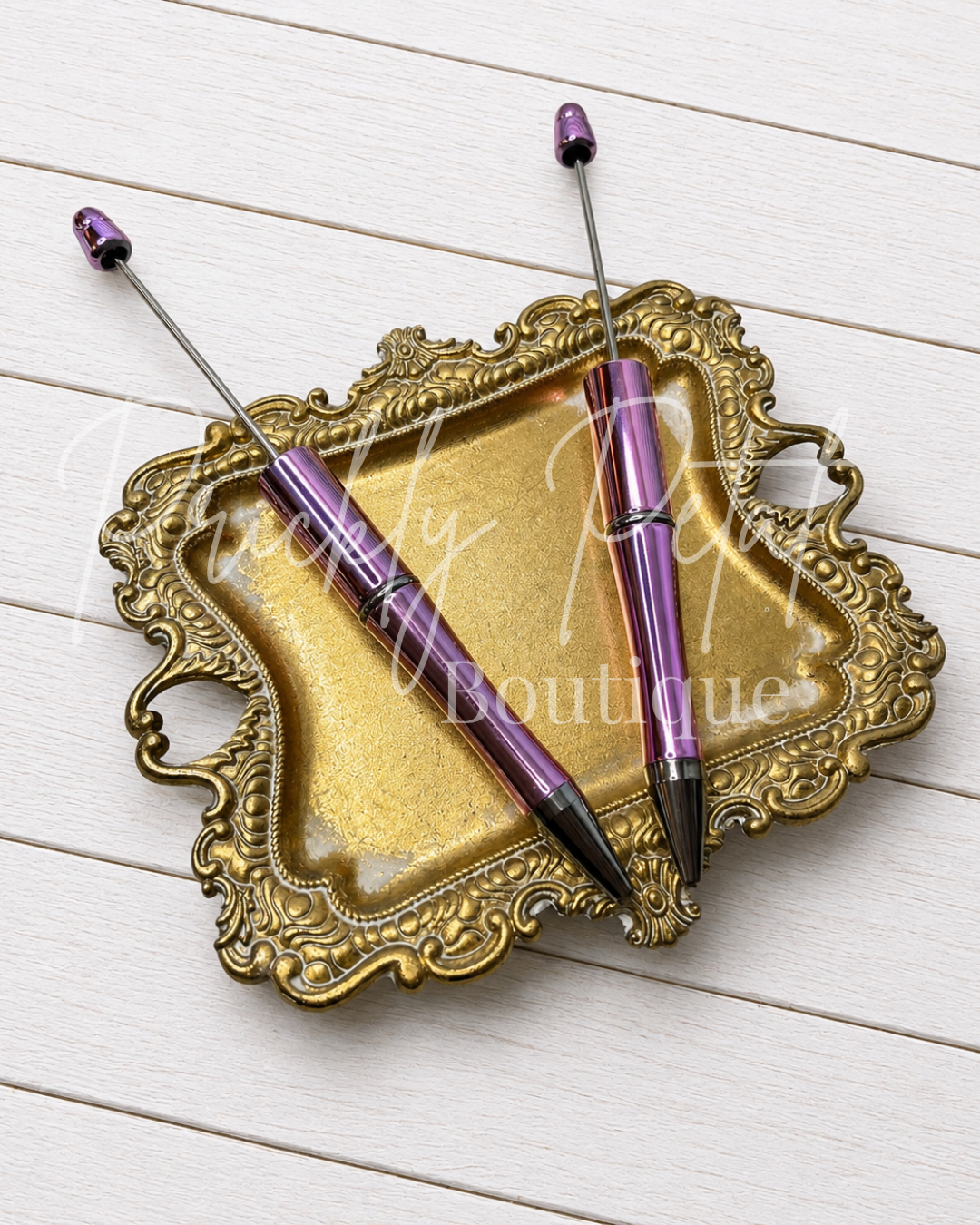 Two metallic purple fountain pens with black tips resting on an ornate gold tray, placed on a white wood plank background. 