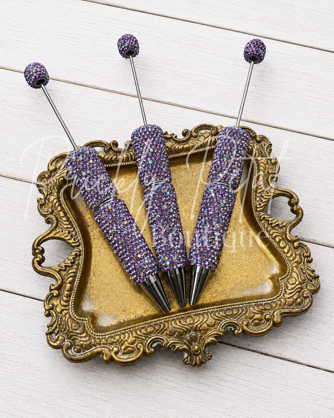 Three purple rhinestone-coveredpens with long metal handles and round purple tips, arranged on an ornate gold tray over a white wood background.