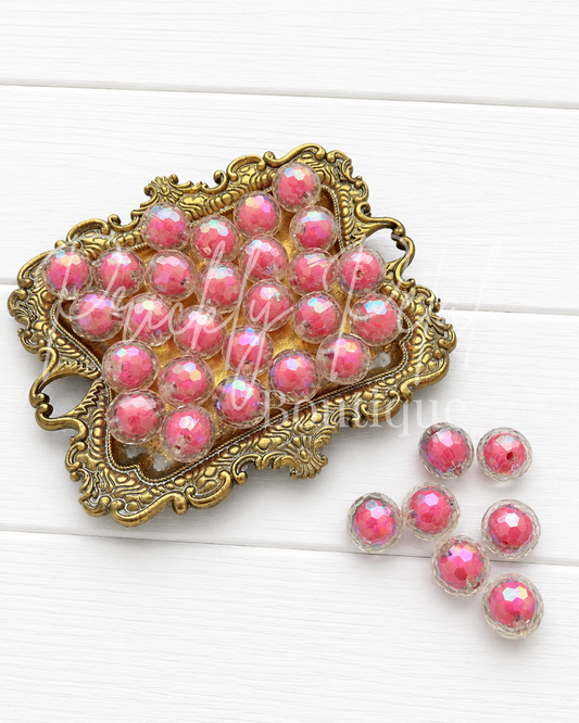 Pink faceted beads with an iridescent finish, displayed inside an ornate gold-colored metal dish with a few loose beads on a white wooden background.