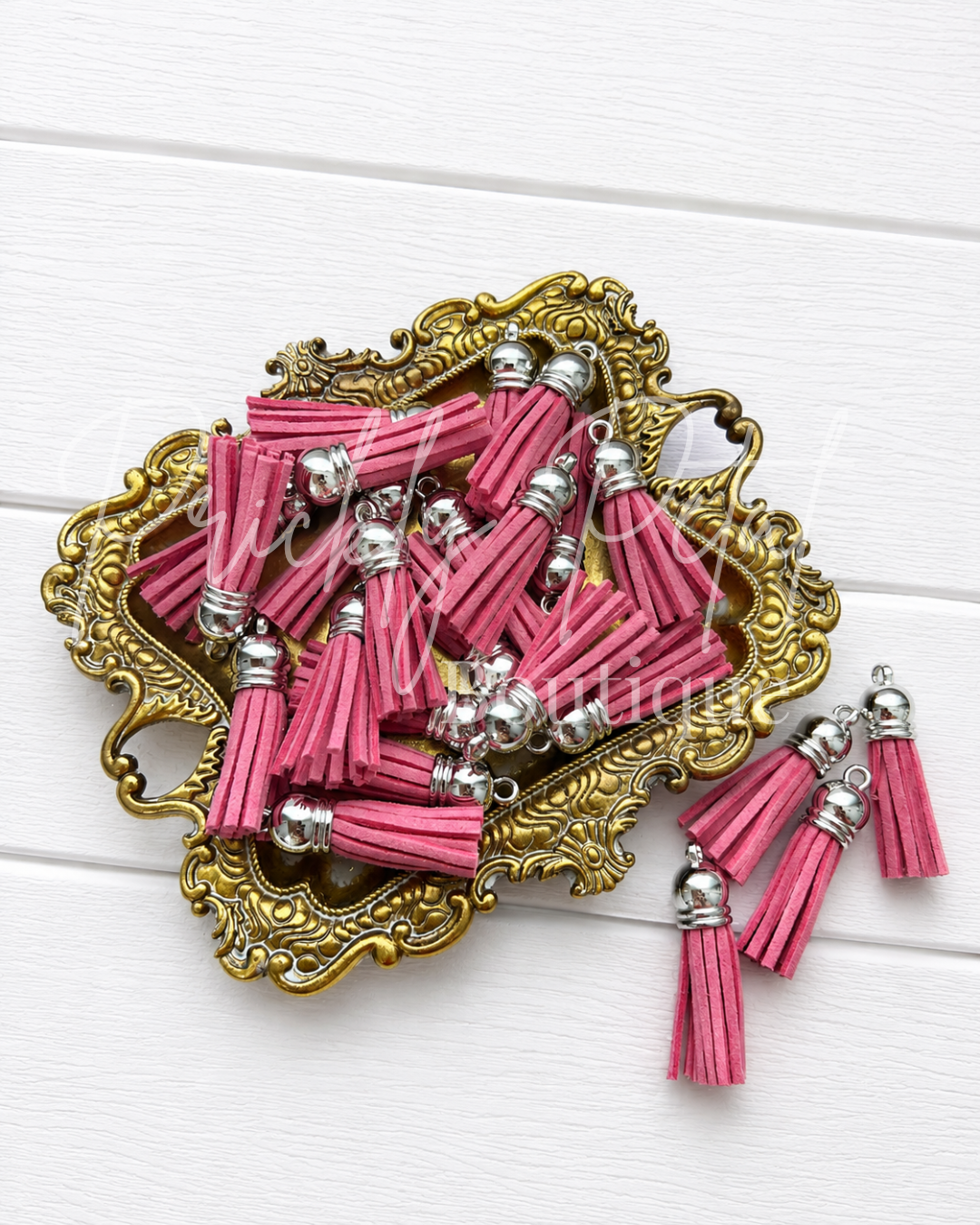 Pink faux leather tassel charms with silver metal caps and loops, displayed on an ornate gold tray against a white wood background.