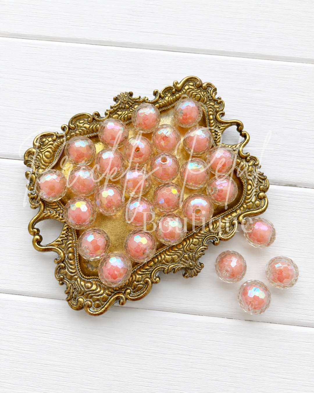A collection of faceted peach-pink iridescent beads displayed on an ornate gold-colored metal tray, with a few beads spilled onto the white wooden surface below. 