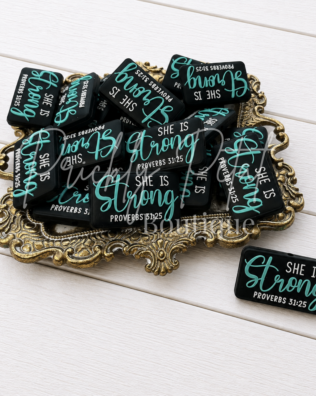 A set of black rectangular magnets with teal and white text reading 'She Is Strong' and 'Proverbs 31:25' arranged on an ornate gold tray over a white wood background.