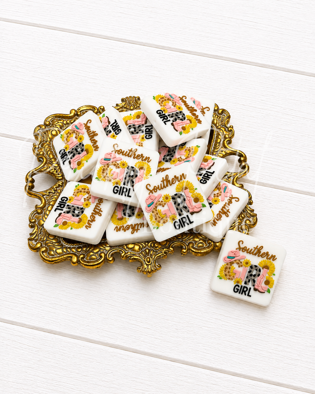 A group of small square white charms with a colorful floral and cow print and the words 'Southern GIRL' in gold and black, displayed on an ornate gold tray over a white wood background.