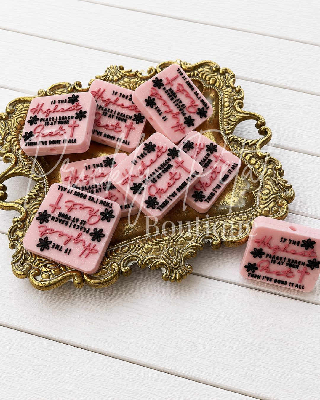 Six pink rectangular beads with black floral and script text designs are arranged on an ornate gold tray, with one soap bar placed beside it on a white wood plank surface.