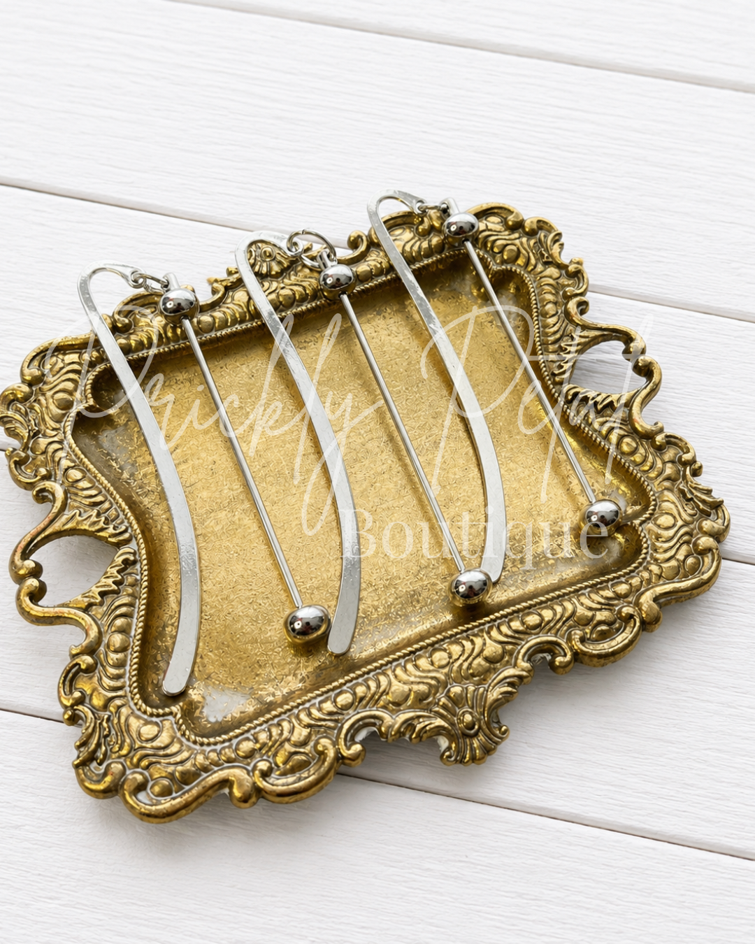 A set of five silver metal beadable bookmark arranged on an ornate, vintage-style gold-colored metal tray, photographed on a white painted wood plank background.