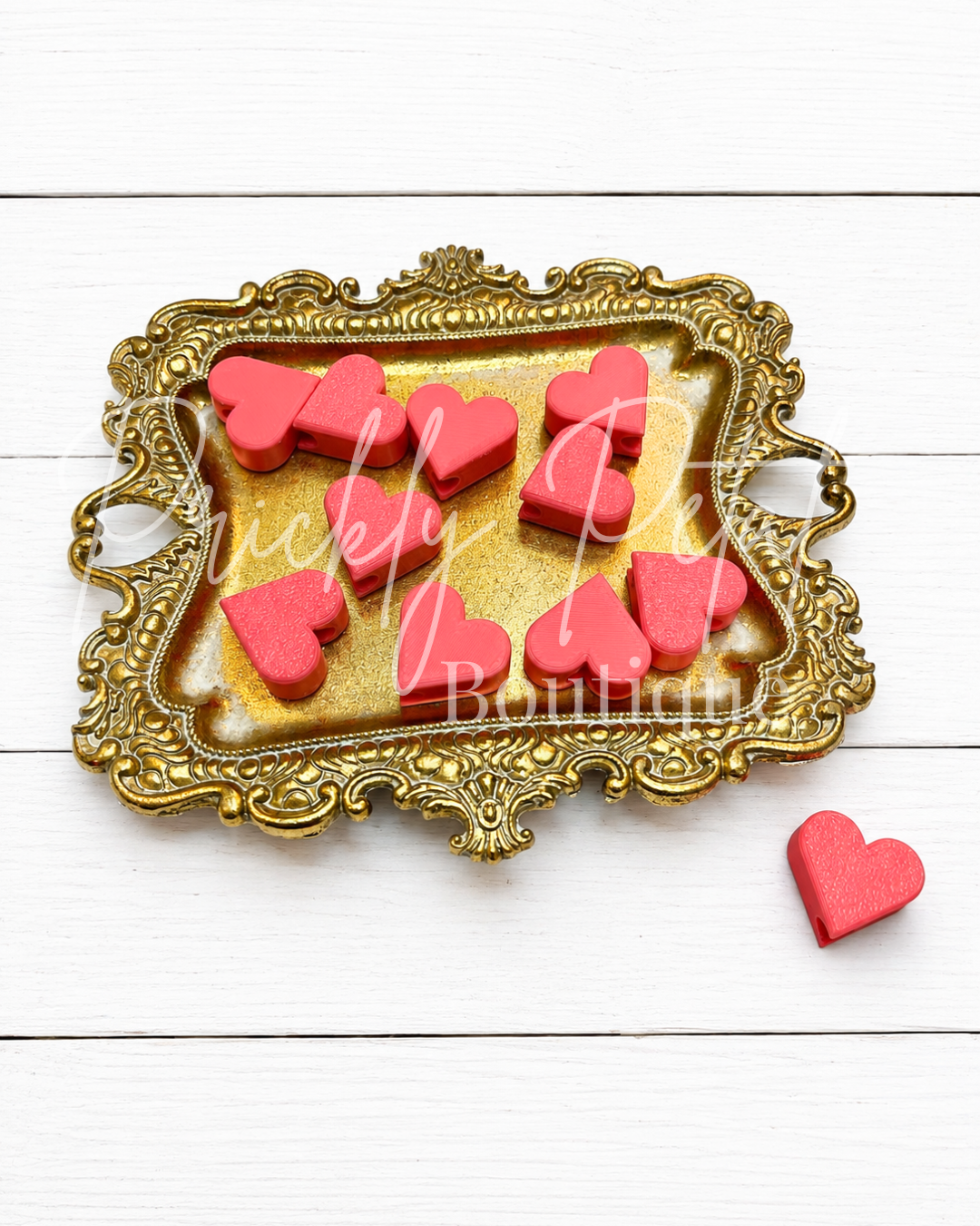 A set of pink heart-shaped beads or pendants displayed on an ornate gold tray, with one matching pink heart placed beside the tray on a white wood plank background. 