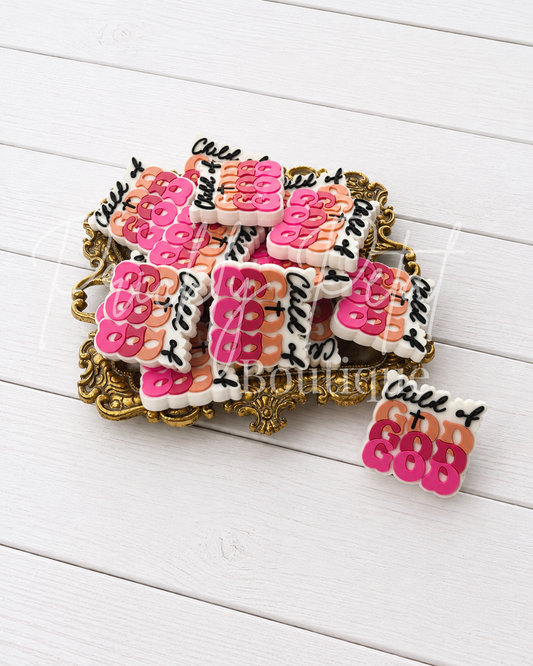 A pile of small rectangular charms or badges with pink, peach, black, and white lettering, displayed on an ornate gold tray over a white wood plank background.