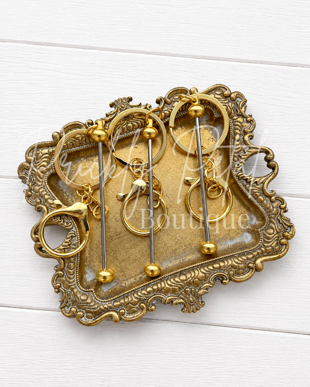 Four gold and silver-toned beadable bar keychains displayed on an ornate vintage-style gold tray against a white wood plank background.