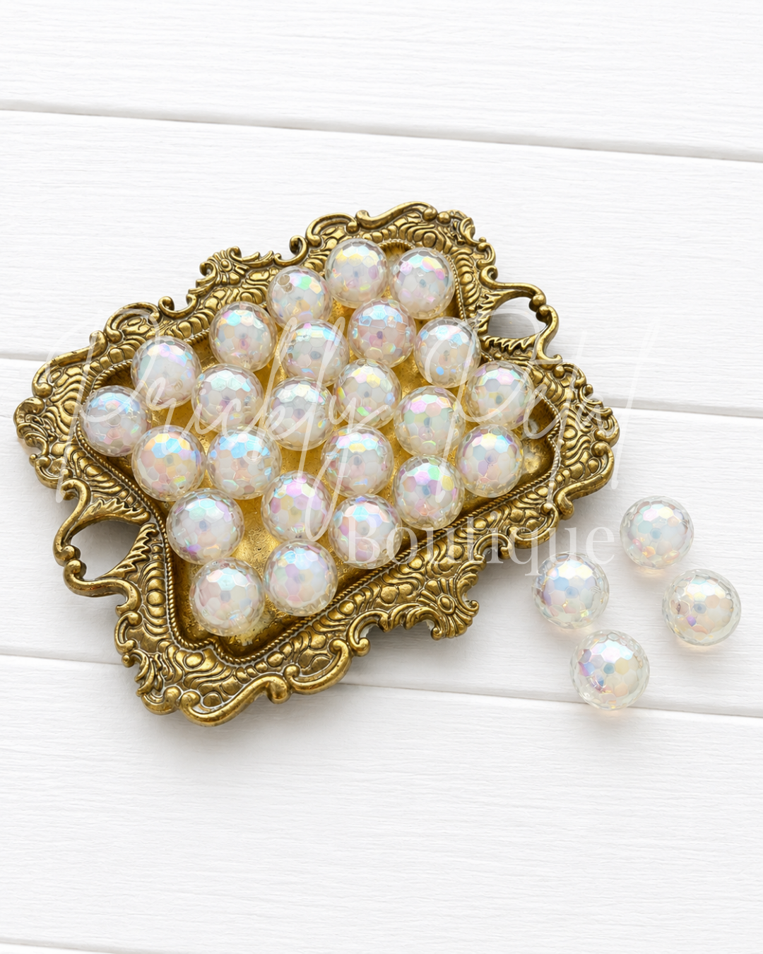 White iridescent pearl beads arranged in an ornate gold metal dish on a white wood background, with three beads placed to the right. 