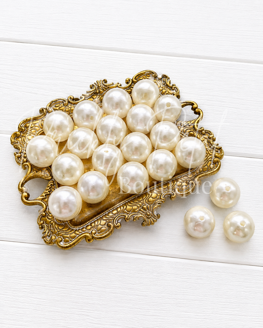 A group of round, glossy white faux pearls arranged on an ornate gold tray, with three additional pearls placed beside it on a white wood background.