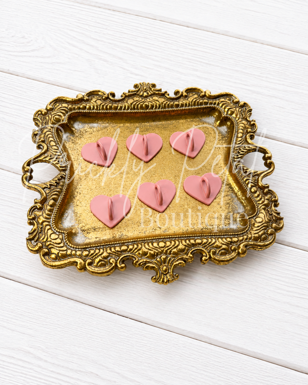 Six pink heart-shaped decorative items arranged on a gold ornate tray with a clear top, placed on a white wood plank background.