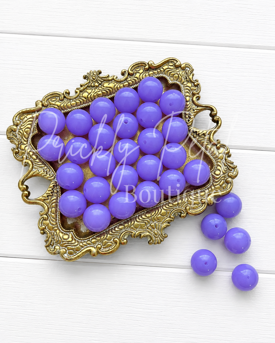 Purple round craft beads arranged in an ornate gold tray on a white wood background, with a few beads spilled beside the tray. 