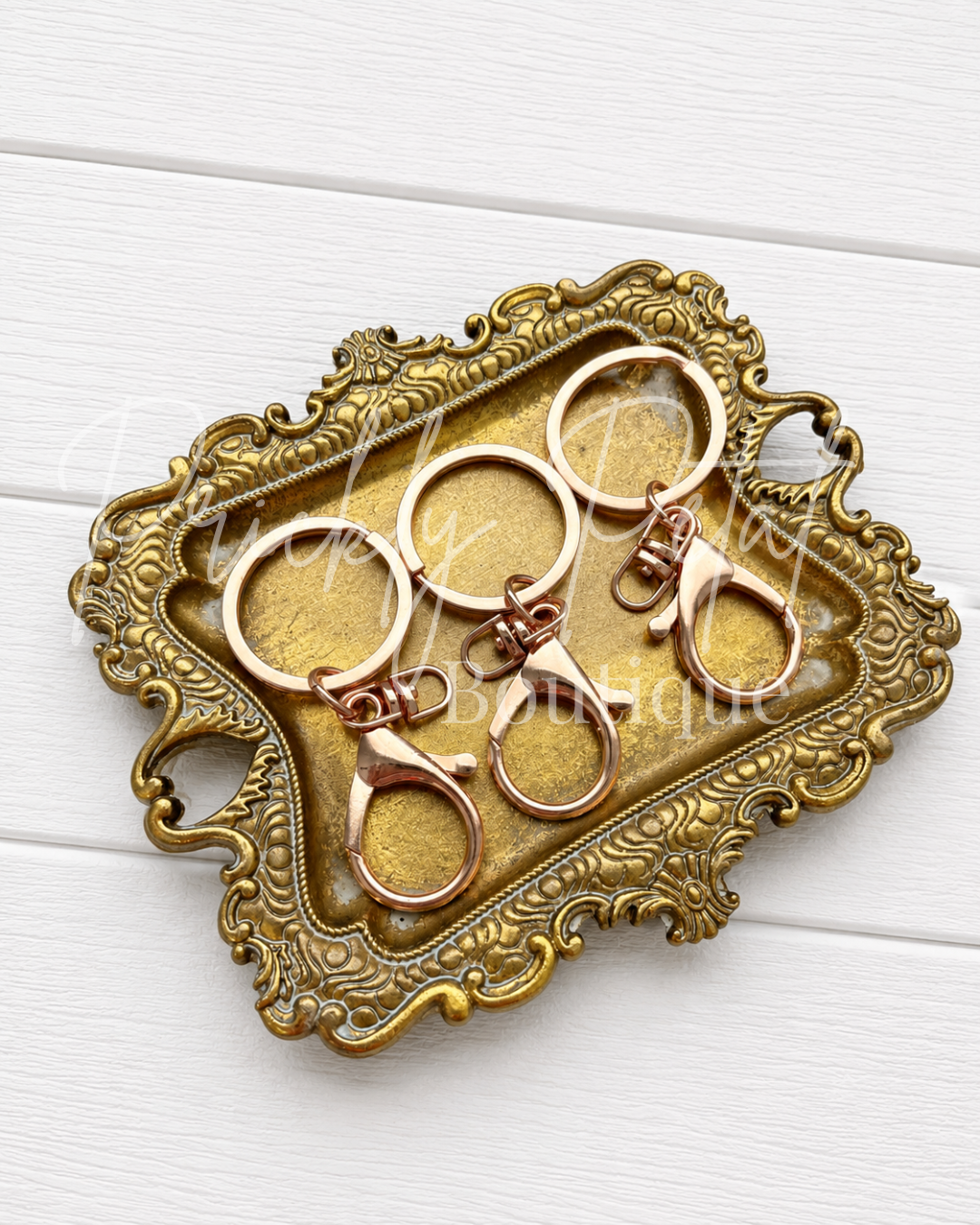 Four rose gold metal key rings with lobster clasps arranged on an ornate vintage-style gold tray over a white wood background.