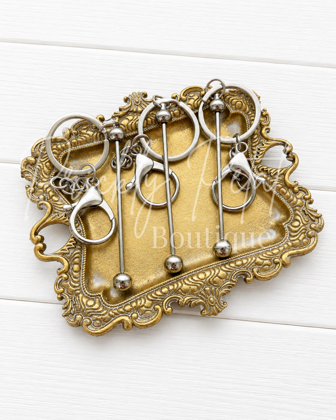 Four silver-toned beadable bar keychains displayed on an ornate gold-colored tray over a white wood background.