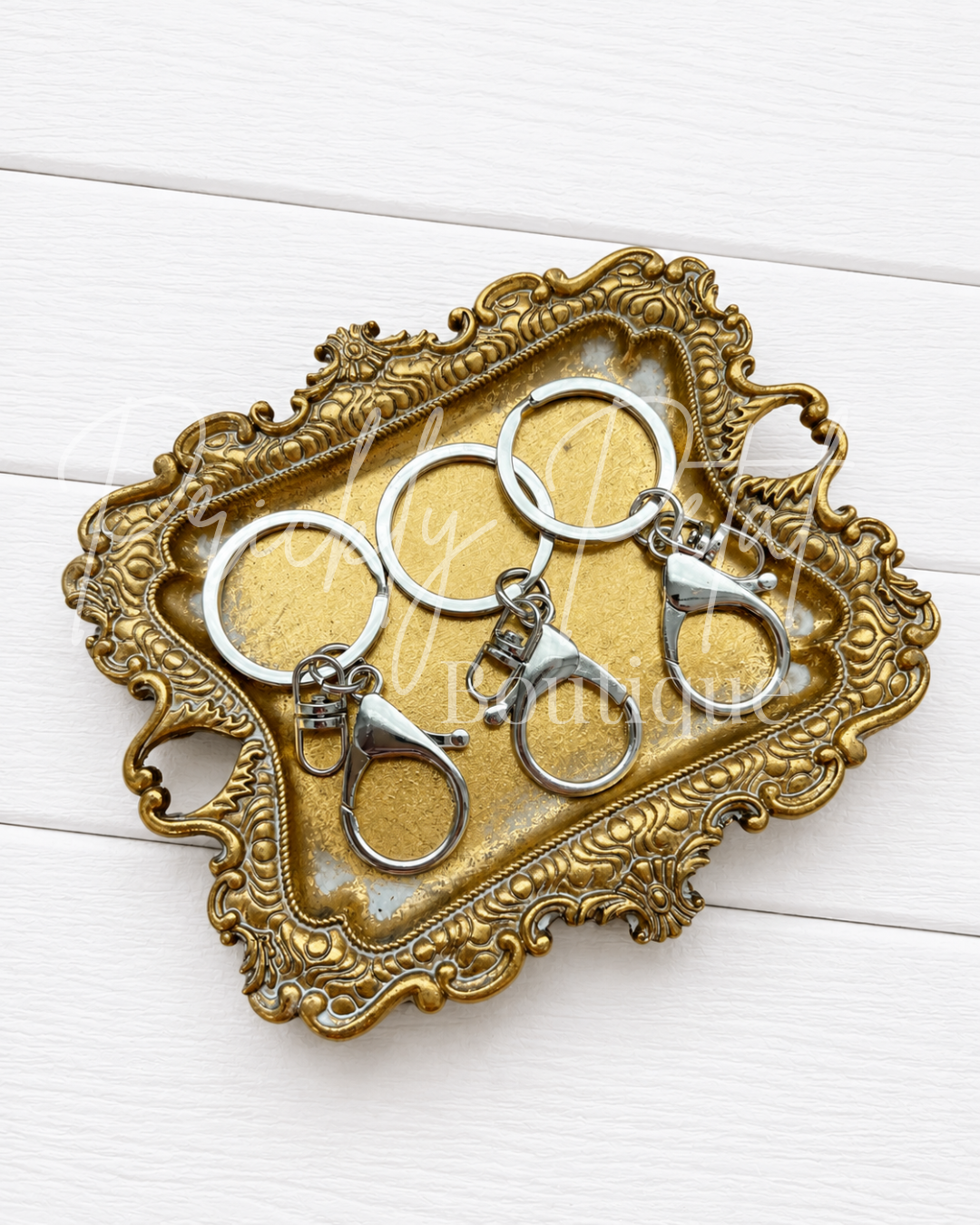 Four silver lobster clasp keychains arranged on an ornate gold-colored tray, set against a white wood plank background.