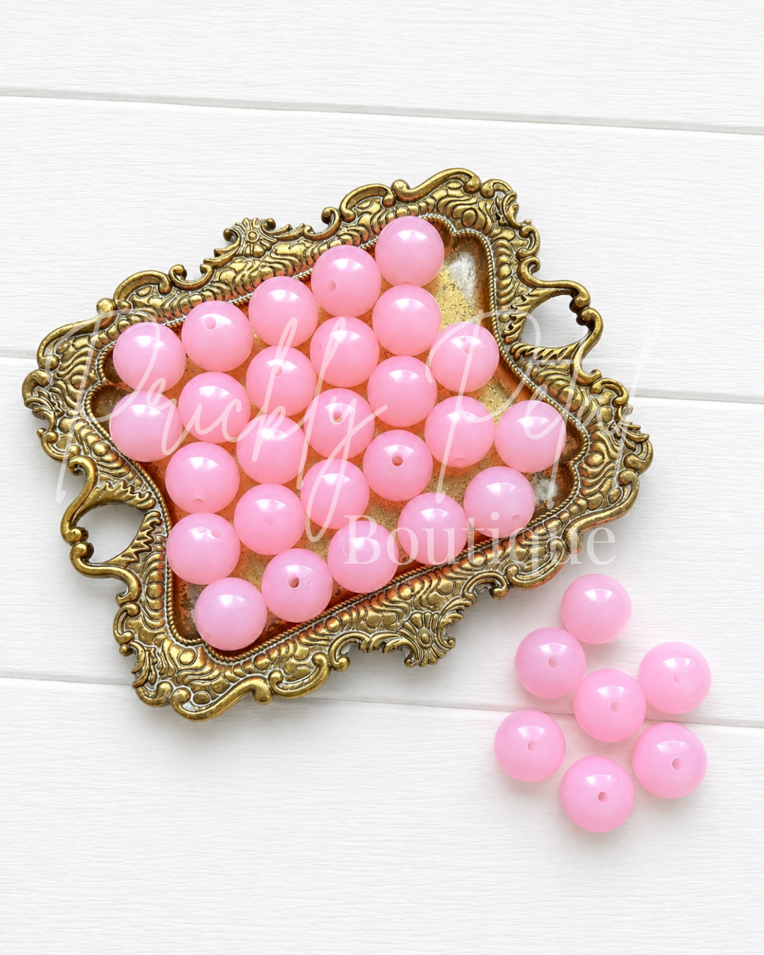 Pink round craft beads arranged on an ornate gold tray, with a few beads placed beside it on a white wood background. 