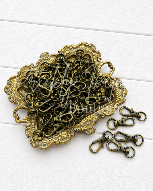 A pile of antique bronze metal lobster clasp swivel snaps arranged on an ornate matching tray, with five clasp snaps displayed separately on the right, set against a white wood plank background.