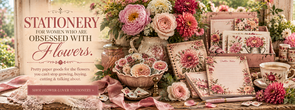A styled tabletop display of floral-themed stationery and paper goods, including cards, envelopes, labels, and decorative paper, arranged with pink flowers, a teacup, and a sign that reads 'Stationery for women who are obsessed with flowers.'.